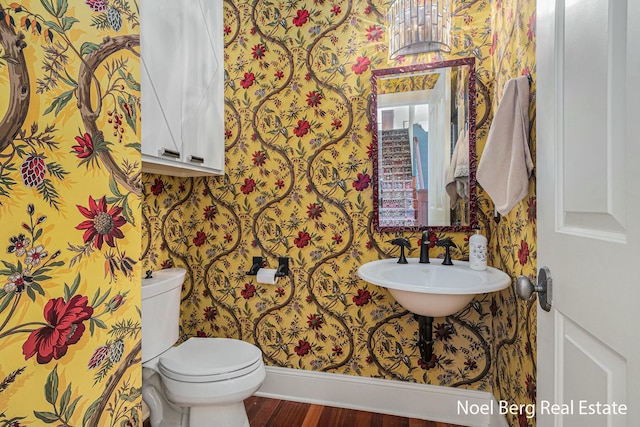 half bathroom featuring wallpapered walls, baseboards, toilet, wood finished floors, and a sink