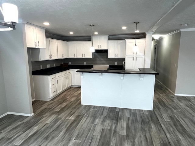 kitchen featuring a kitchen island with sink, dark wood-style flooring, under cabinet range hood, dark countertops, and backsplash