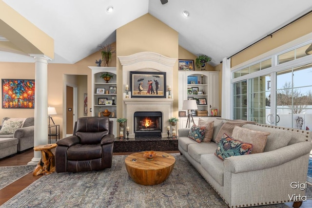 living room featuring high vaulted ceiling, a warm lit fireplace, wood finished floors, and ornate columns