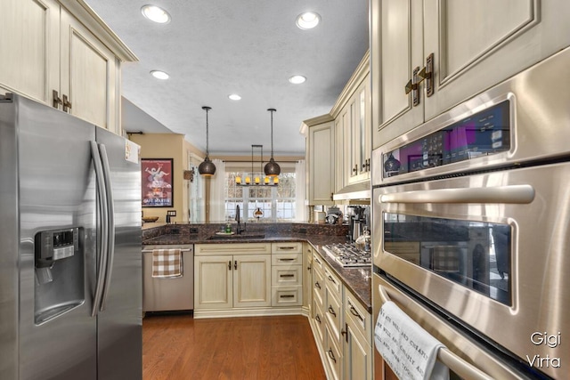 kitchen with a sink, decorative light fixtures, appliances with stainless steel finishes, cream cabinetry, and dark wood-style flooring