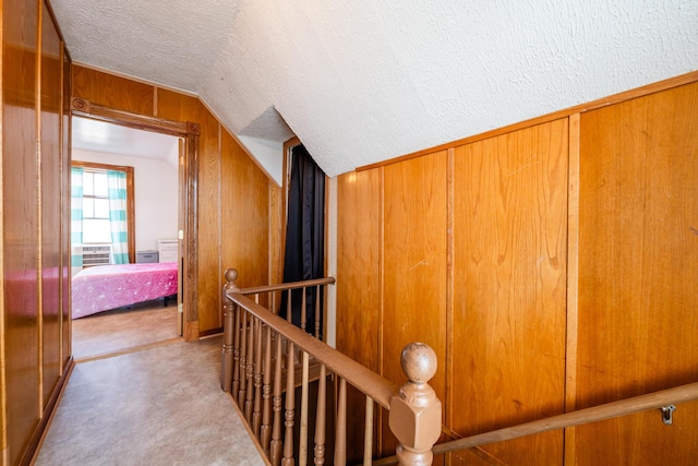 hallway featuring wooden walls, carpet floors, an upstairs landing, vaulted ceiling, and a textured ceiling