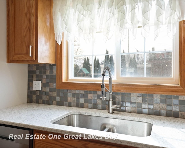 kitchen featuring dishwasher, light stone counters, decorative backsplash, brown cabinetry, and a sink