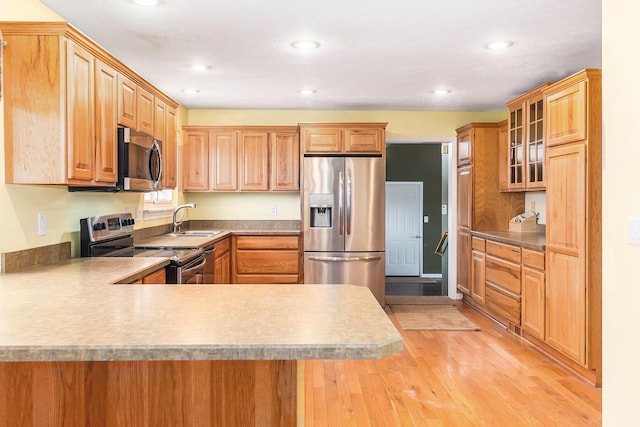 kitchen featuring a sink, appliances with stainless steel finishes, a peninsula, light wood finished floors, and glass insert cabinets