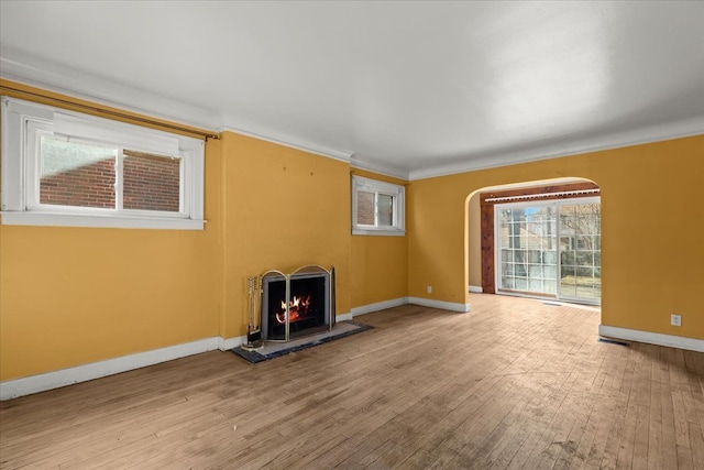 unfurnished living room with baseboards, arched walkways, a warm lit fireplace, wood-type flooring, and crown molding