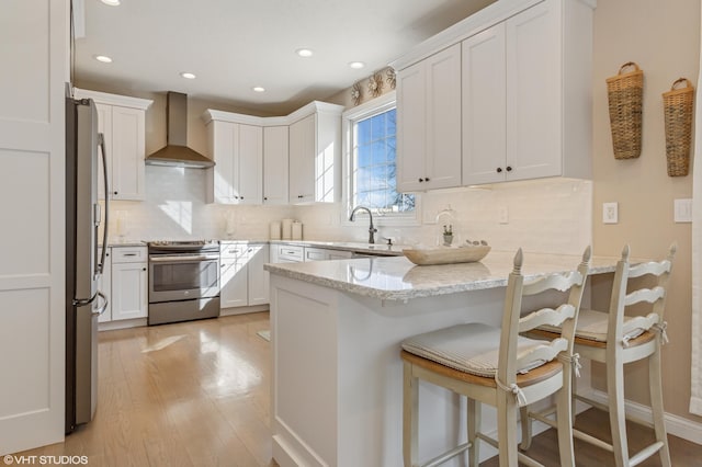 kitchen featuring a sink, stainless steel appliances, a peninsula, wall chimney exhaust hood, and light stone countertops