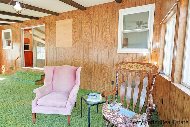 sitting room with a ceiling fan, wooden walls, carpet, and beamed ceiling