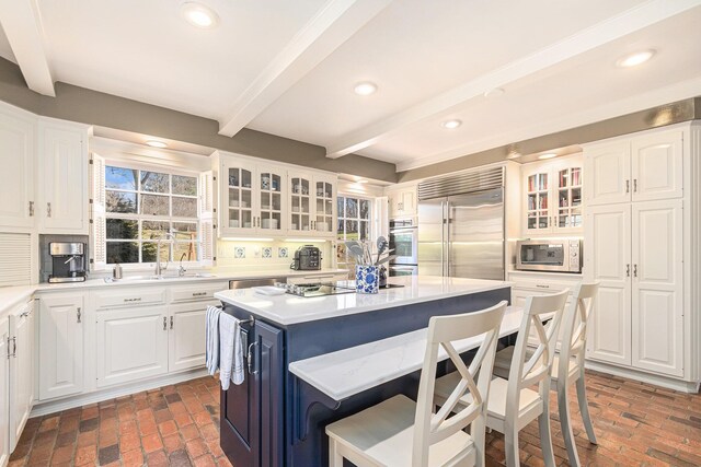 kitchen featuring a kitchen breakfast bar, appliances with stainless steel finishes, beam ceiling, a sink, and brick floor