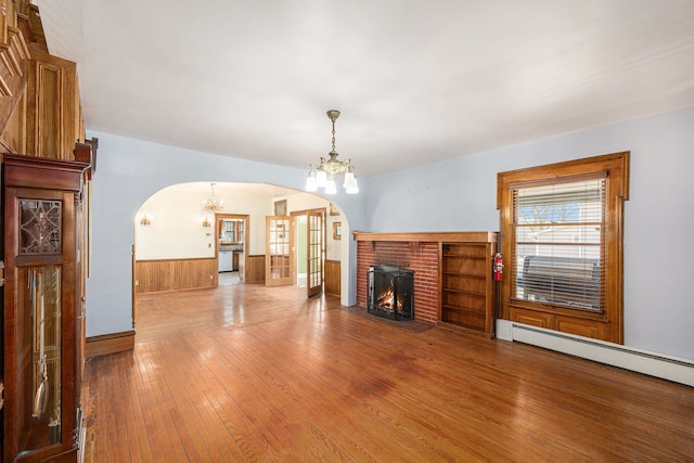 unfurnished living room featuring arched walkways, wainscoting, a brick fireplace, a notable chandelier, and baseboard heating