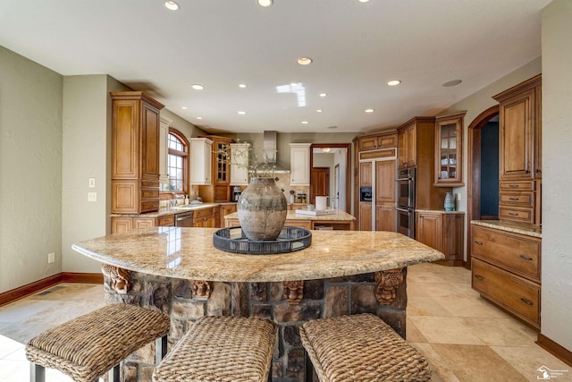 kitchen with a breakfast bar, a spacious island, brown cabinetry, wall chimney range hood, and glass insert cabinets