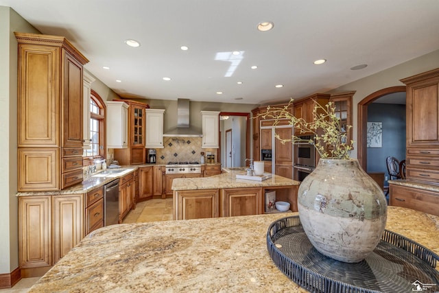 kitchen with backsplash, light stone counters, stainless steel appliances, wall chimney exhaust hood, and a sink