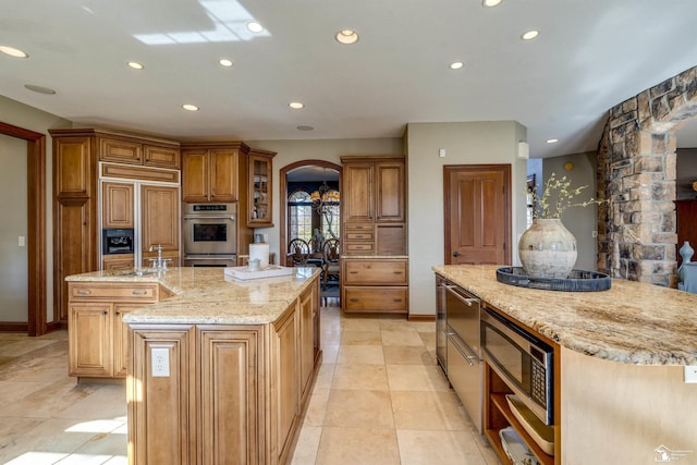 kitchen with recessed lighting, arched walkways, a kitchen island with sink, double oven, and brown cabinets