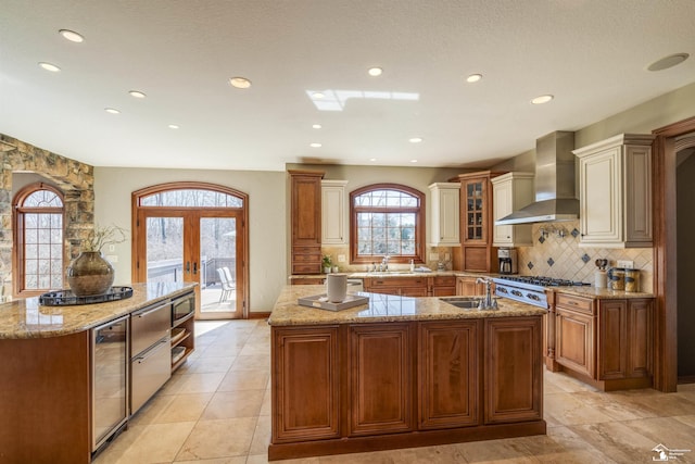 kitchen with backsplash, an island with sink, stainless steel gas stovetop, wall chimney exhaust hood, and a sink