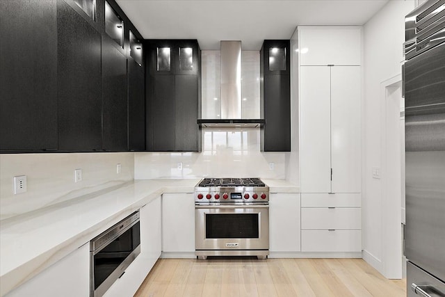 kitchen with dark cabinetry, light wood-style floors, appliances with stainless steel finishes, wall chimney exhaust hood, and light countertops