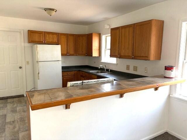 kitchen with a peninsula, freestanding refrigerator, a sink, a kitchen bar, and brown cabinets