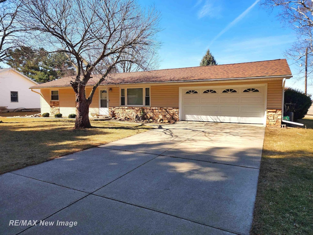 ranch-style house featuring an attached garage, a shingled roof, concrete driveway, a front lawn, and stone siding