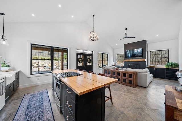 kitchen with butcher block countertops, ceiling fan with notable chandelier, stainless steel range with gas stovetop, a fireplace, and dark cabinets