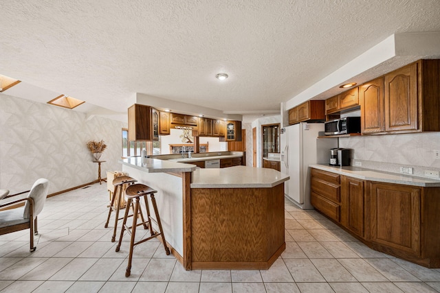 kitchen featuring a breakfast bar, white appliances, a peninsula, brown cabinetry, and wallpapered walls