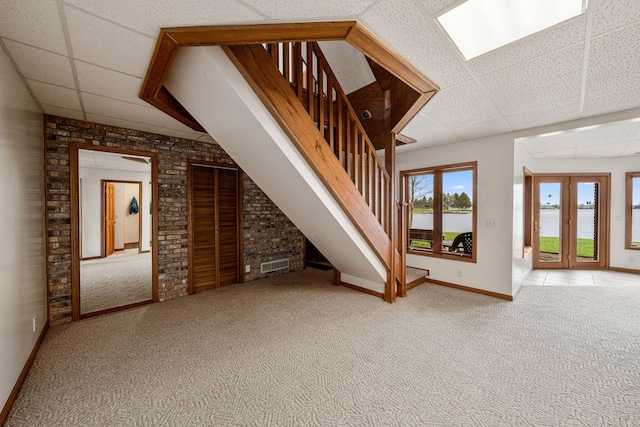 unfurnished living room featuring visible vents, carpet floors, brick wall, a paneled ceiling, and stairs