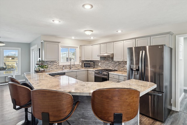 kitchen featuring a sink, under cabinet range hood, backsplash, appliances with stainless steel finishes, and a peninsula