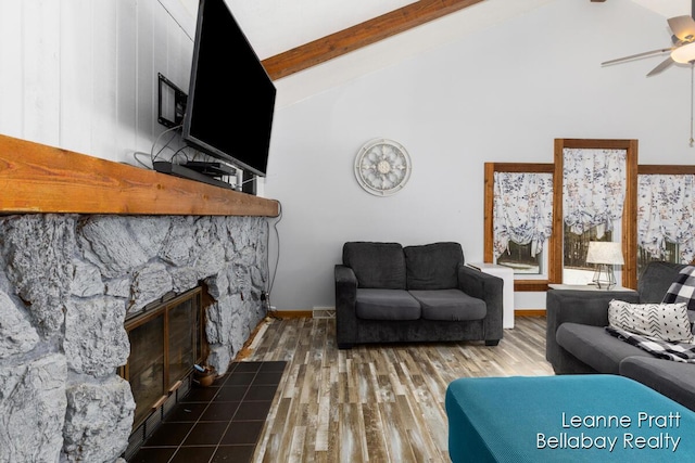 living area featuring beam ceiling, high vaulted ceiling, a ceiling fan, wood finished floors, and a stone fireplace