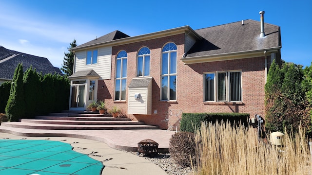 back of property with a covered pool, a patio, a fire pit, roof with shingles, and brick siding