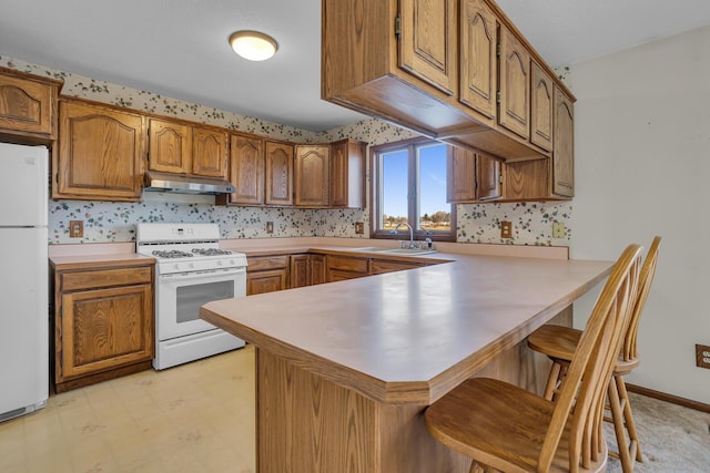 kitchen featuring wallpapered walls, under cabinet range hood, a peninsula, white appliances, and a sink