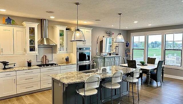 kitchen featuring light wood finished floors, tasteful backsplash, wall chimney range hood, stainless steel double oven, and white cabinetry