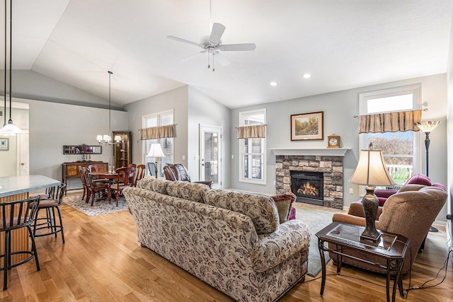 living area featuring a stone fireplace, light wood-style flooring, ceiling fan with notable chandelier, and lofted ceiling