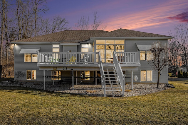 back of house at dusk featuring a lawn, a deck, stairs, and a shingled roof