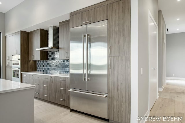 kitchen featuring wall chimney range hood, tasteful backsplash, light wood-type flooring, and appliances with stainless steel finishes