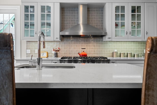 kitchen with backsplash, glass insert cabinets, white cabinets, wall chimney exhaust hood, and a sink