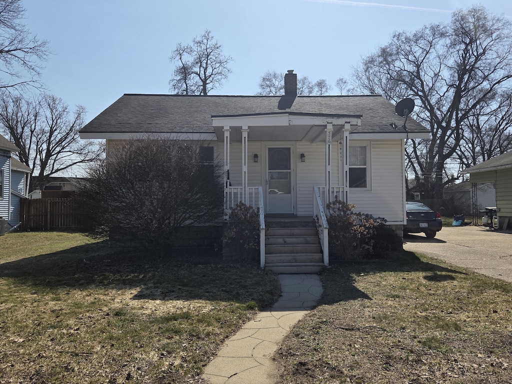 bungalow with a porch, fence, a front yard, a shingled roof, and a chimney