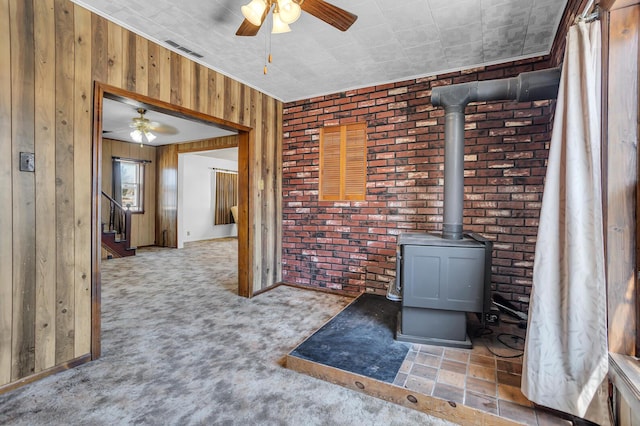 unfurnished living room with wooden walls, visible vents, brick wall, ceiling fan, and a wood stove