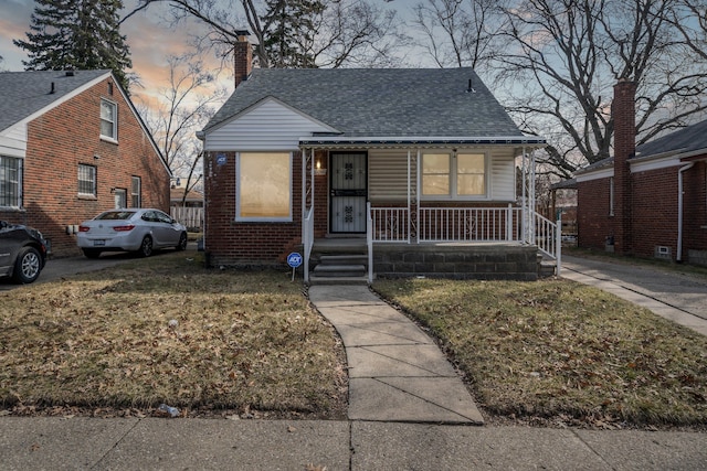 bungalow-style home with brick siding, a shingled roof, covered porch, a chimney, and a yard