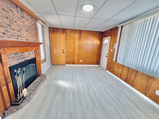 unfurnished living room featuring wooden walls, a brick fireplace, a paneled ceiling, and light wood-style floors