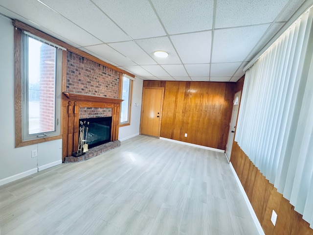 unfurnished living room with baseboards, a drop ceiling, wood walls, light wood-style flooring, and a fireplace