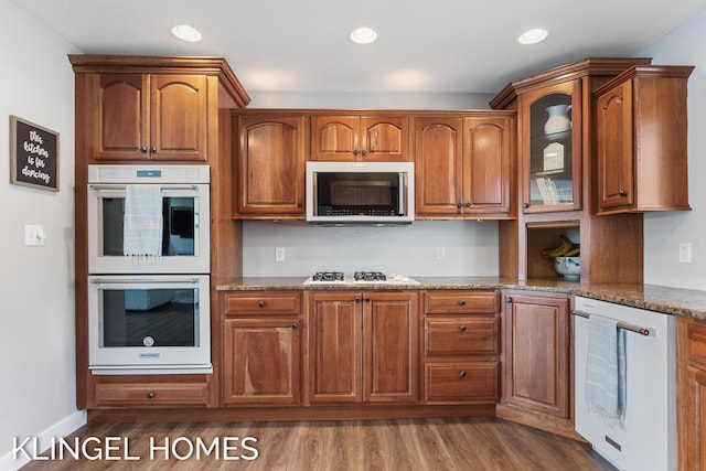 kitchen with white appliances, stone counters, dark wood-type flooring, glass insert cabinets, and brown cabinets