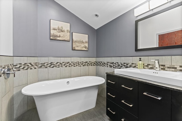 bathroom with tile walls, vaulted ceiling, wainscoting, a freestanding tub, and vanity