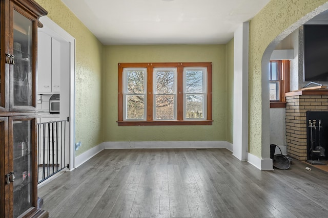 unfurnished living room with wood-type flooring, arched walkways, baseboards, a brick fireplace, and a textured wall