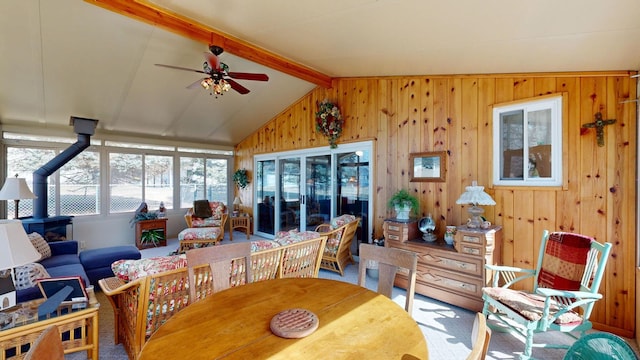 carpeted dining space with lofted ceiling with beams, a wood stove, wooden walls, and ceiling fan