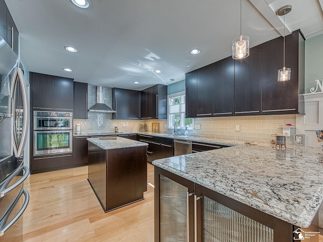 kitchen featuring appliances with stainless steel finishes, wall chimney exhaust hood, light wood finished floors, decorative backsplash, and light stone countertops