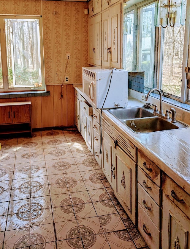 kitchen with wallpapered walls, a wainscoted wall, light countertops, light tile patterned floors, and a sink