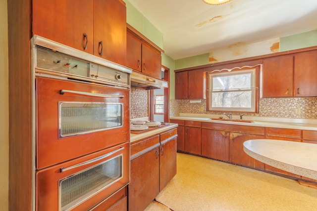 kitchen featuring double wall oven, a sink, light countertops, under cabinet range hood, and tasteful backsplash