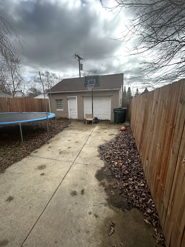 back of property with a trampoline, a fenced backyard, an outdoor structure, driveway, and a patio