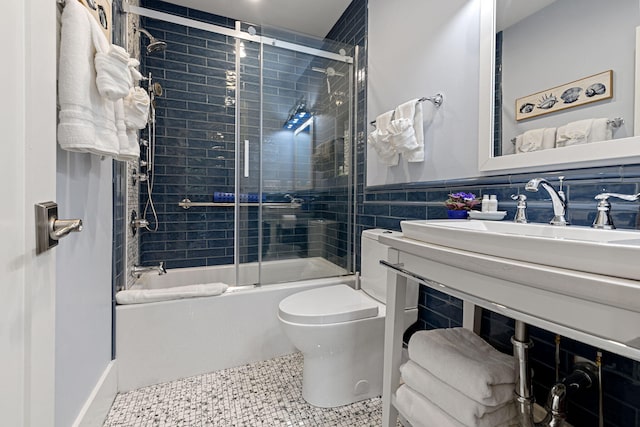 bathroom featuring a wainscoted wall, toilet, a sink, shower / bath combination with glass door, and tile walls