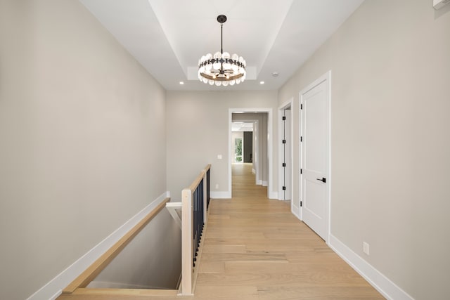 hallway featuring light wood-type flooring, an upstairs landing, a raised ceiling, a notable chandelier, and baseboards