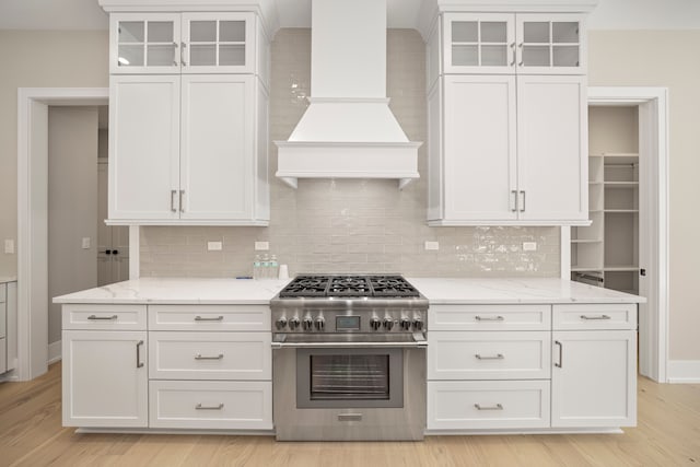 kitchen with premium range hood, stainless steel range, light stone counters, white cabinetry, and light wood finished floors