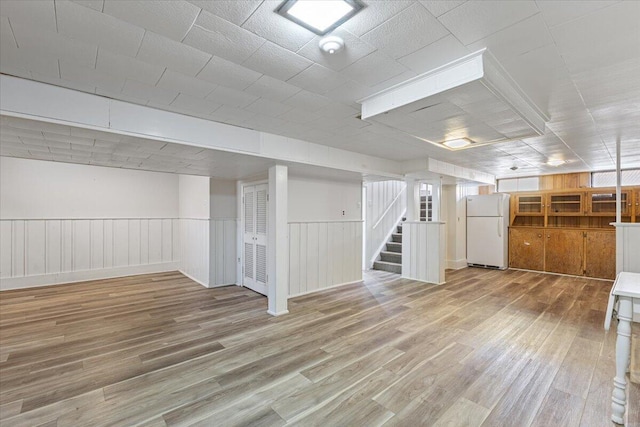 basement with light wood-type flooring, a wainscoted wall, stairs, and freestanding refrigerator