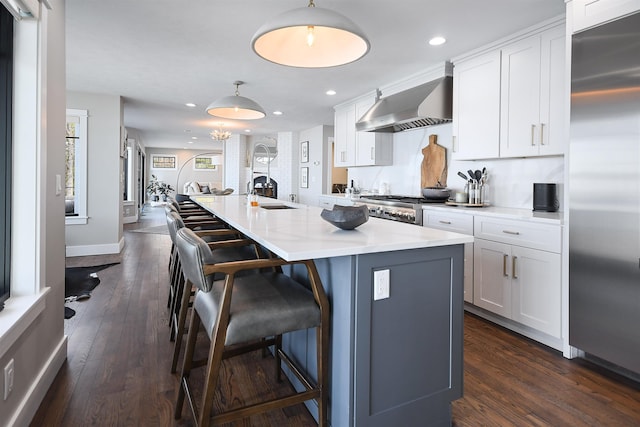 kitchen with a sink, high end refrigerator, a breakfast bar area, wall chimney exhaust hood, and dark wood-style flooring