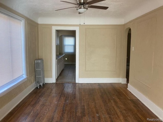unfurnished room with arched walkways, dark wood-style flooring, a ceiling fan, and radiator heating unit